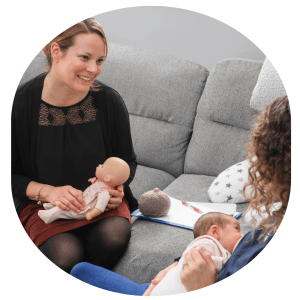 Liz Farrant sitting on a grey sofa and holding a doll, observing a new Mummy breastfeeding her newborn baby