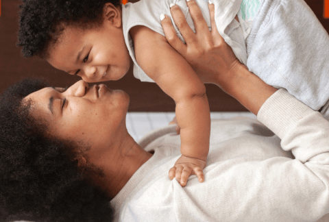 Mother laying on bed and holding baby above her, kissing baby on the cheek. Both smiling