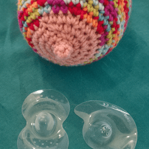 Knitted breast in vibrant colours sitting on a green background. 2 nipple shields in front of the knitted breast